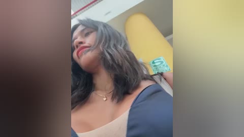 Media: Video of a light-skinned woman with shoulder-length dark hair, wearing a black and beige top, red lipstick, and a gold necklace, standing in a yellow and white room.