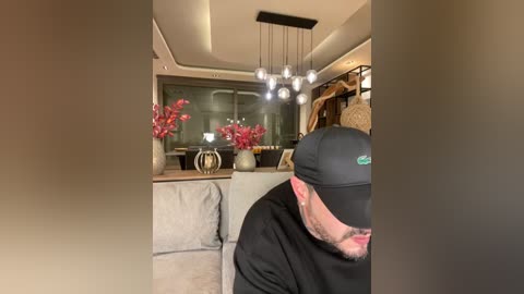 Media: Video of a man in a black cap and jacket, facing away, in a modern, warmly lit living room with gray sofa, red flowers, and minimalist decor.