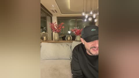 Media: Video of a man with a beard and black baseball cap smiling, sitting on a beige sofa in a modern living room with mirrored walls, a glass coffee table, and decorative vases.