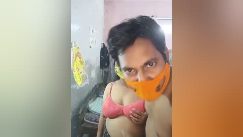 Media: A video of a woman with medium skin tone, wearing a pink bra and a yellow face mask, standing in a dimly lit room with yellow caution tape on a wall.