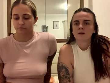 Media: Video of two women seated in a bathroom. One has light skin, long blonde hair, wearing a pink top. The other has dark skin, long dark hair, tattoos, and wears a white sleeveless top.