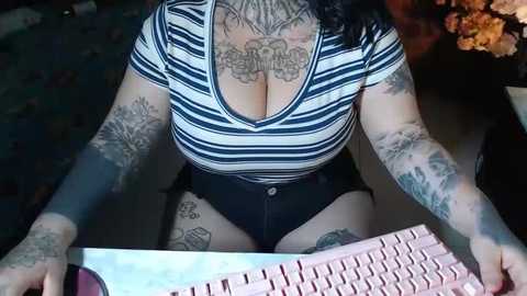 Media: Video of a tattooed, curvy woman with large breasts, wearing a striped top, sitting at a computer, with a pink keyboard in focus.