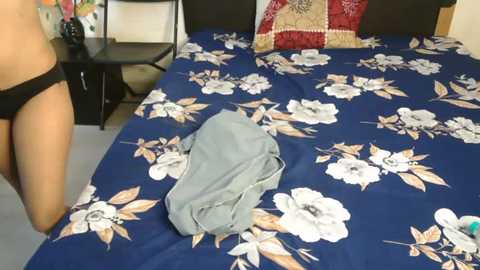 Media: A video showing a woman wearing black underwear on a bed with a floral blue blanket, discarded light grey shirt, and red pillow in a modern bedroom.