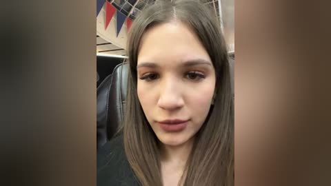 Media: A video of a young Caucasian woman with light skin and long, straight brown hair. She has a neutral expression and is sitting in a bus with a festive, colorful pennant banner in the background.