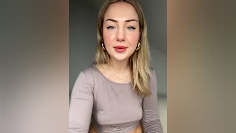 Media: Video of a fair-skinned, blonde woman with light makeup, wearing a long-sleeved, light pink top, against a blurred background.