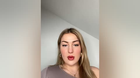 Media: Video of a young woman with fair skin, straight blonde hair, and full lips, wearing a light grey off-shoulder top. She stands against a plain white wall, with a soft, neutral background.