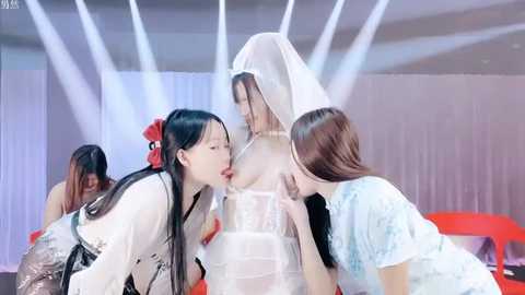 Media: Video: Two Asian women in bridal gowns, one with a veil, kissing passionately. Background features modern, white-lit stage with red chairs.