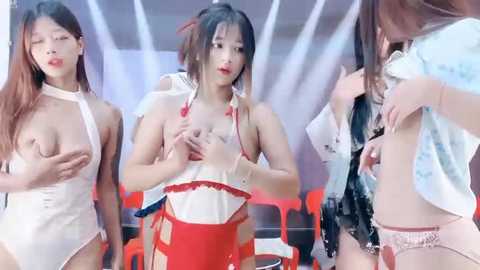 Media: Video of three Asian women in revealing costumes, one with exposed breasts, another partially undressed, and the third wearing a revealing red dress. Background shows modern interior with red chairs and white walls.