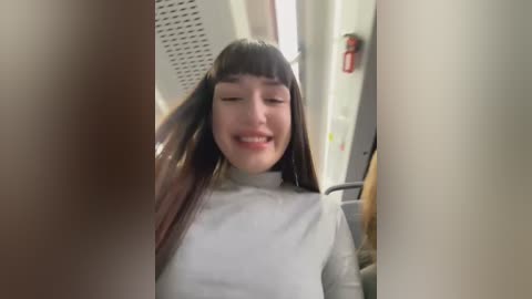 Media: Video of an Asian woman with long, straight, dark hair, wearing a light gray turtleneck, smiling in a train compartment. The background shows beige walls, a fire extinguisher, and luggage.