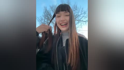 Media: Video of a young Asian woman with long, straight, black hair with orange highlights, wearing a black leather jacket, smiling and playfully pulling a strand of her hair. Background features a clear blue sky and leafless trees.