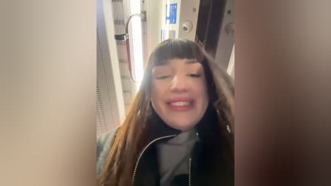 Media: A video of a smiling woman with long, straight brown hair, wearing a black jacket, standing in a narrow, brightly lit subway car.