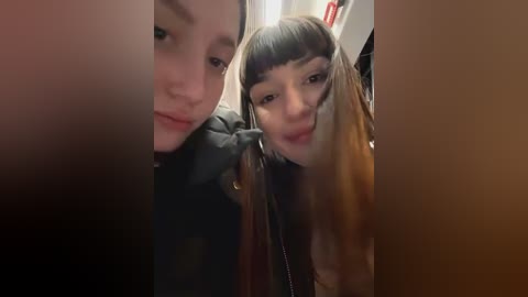 Media: Video of two young women, one with blunt bangs, another with long, straight hair, both smiling and making silly faces, wearing dark coats, indoors.