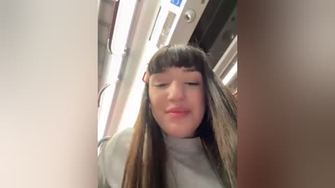 Media: Video of a young woman with straight, long brown hair, wearing a white turtleneck, looking down, inside a dimly lit, narrow subway car.