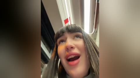 Media: Video of a woman with long, straight gray hair, wearing a gray coat, captured from a low angle inside a train, mouth open in a joyful expression, surrounded by beige and brown interior.