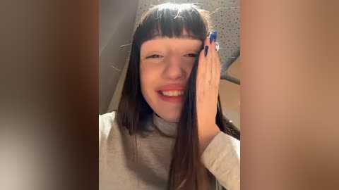 Media: A video of a smiling woman with long brown hair, light skin, and a grey sweater, holding her face with a blue-painted fingernail, set in a dimly lit room.