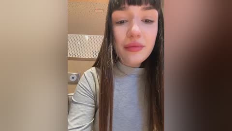 Media: Video of a young woman with long, straight, dark brown hair and blunt bangs, wearing a light gray long-sleeve shirt. She has a neutral expression, standing in a bathroom with beige walls, a tiled backsplash, and a roll of toilet paper.