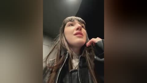 Media: Video of a young woman with long brown hair, wearing a black jacket, touching her chin, in a dimly lit, narrow hallway with a ceiling light.