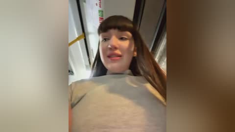 Media: Video of a young woman with straight brown hair, fair skin, and light makeup, wearing a beige top. She's in a train, with a blurred background of white and yellow signs.