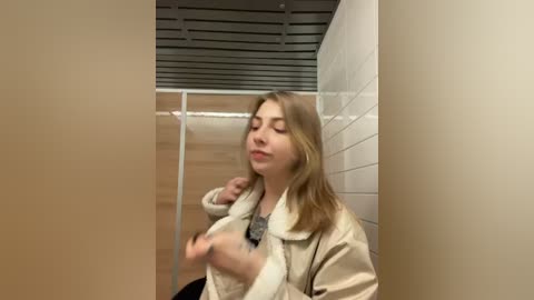 Media: Video of a young Caucasian woman with shoulder-length blonde hair, wearing a beige coat, standing in a narrow, tiled bathroom with white walls and a metal door.