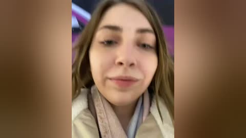 Media: Video of a young woman with straight, light brown hair, fair skin, and light makeup. She wears a beige jacket over a light-colored sweater. The background is blurred, likely indoor with a hint of pink and purple.