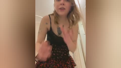 Media: A video of a young woman with fair skin and shoulder-length blonde hair, wearing a black camisole and floral skirt. She has tattoos on her arms and is caught mid-movement, her hand raised defensively. The background is a plain, beige wall.