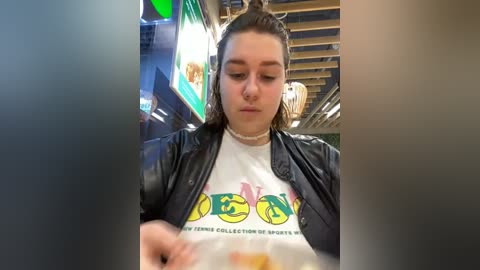 Media: Video of a young woman with long hair in a bun, wearing a white T-shirt with colorful tennis-themed graphics and a black jacket, eating at a restaurant with neon lights and modern decor.