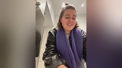 Media: Video of a young woman with shoulder-length blonde hair, smiling, wearing a black leather jacket and a purple towel around her neck, standing in a modern, white-walled restroom.