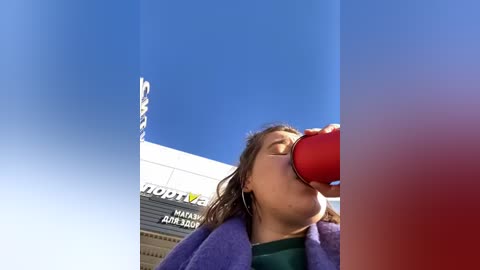 Media: A young woman with long brown hair drinks from a red cup under a clear blue sky. She wears a purple jacket and green shirt. A shopping mall sign is visible in the background.