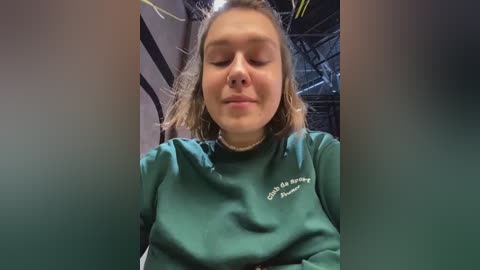 Media: Video of a young woman with light brown hair, wearing a green sweatshirt with a white logo, sitting in a dimly lit room with industrial equipment in the background.