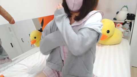 Media: Video of an Asian woman with a large chest, wearing a pink mask, light gray hoodie, and pink shorts, hugging a yellow duck plushie.