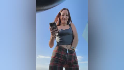 Media: Video of a young woman with light skin and long brown hair, wearing a grey crop top and red plaid pants, smiling and taking a selfie with a smartphone, reflected in a curved mirror.