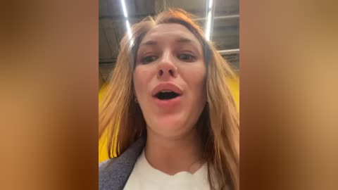 Media: A close-up video of a Caucasian woman with long, light brown hair and fair skin, wearing a white shirt, smiling with her mouth open. Background features an industrial setting with metal beams and yellow lighting.