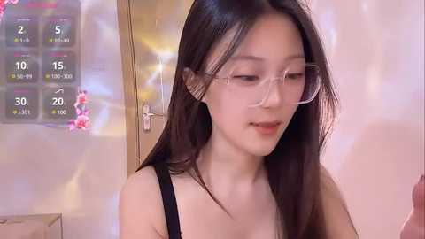 Media: A video of an East Asian woman with long black hair, wearing clear glasses, a black top, and a slight smile. The background shows a calendar and a blurred, softly lit room.