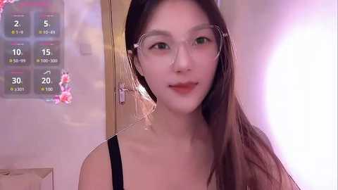 Media: Video of an East Asian woman with long brown hair and glasses, wearing a black top, standing in a softly lit room with a temperature display on the left.