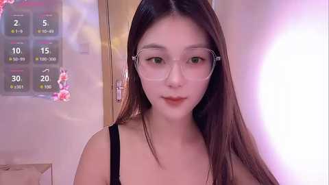 Media: Video of an East Asian woman with long brown hair, wearing clear glasses, a black tank top, and a neutral expression, standing in a room with a digital weather display and soft lighting.