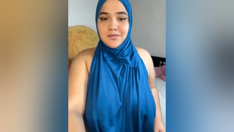 Media: Video of a young, light-skinned woman with medium-length hair, wearing a vibrant blue hijab and sleeveless top, standing in a softly lit bedroom with a yellow bedspread in the background.