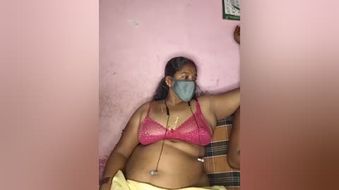 Media: A video of a middle-aged, dark-skinned woman with medium build, wearing a pink lace bra, teal face mask, and yellow towel, seated in a corner of a pink-walled room.