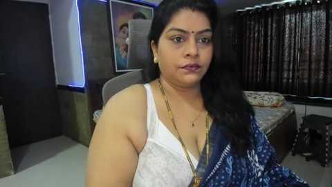 Media: Video of a South Asian woman with medium skin tone, long black hair, wearing a white bra, blue patterned saree, gold necklace, in a modern bedroom with a bed, curtains, and framed photos.