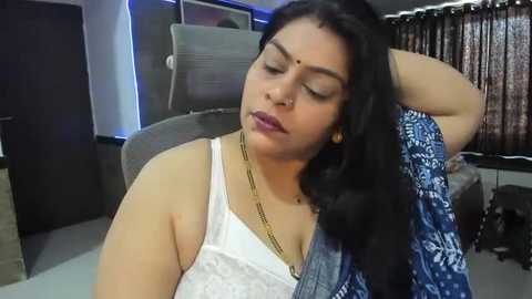 Media: Video of a middle-aged South Asian woman with medium skin tone, wearing a white lace bra and blue patterned saree, leaning back on a gray chair, looking tired. Background shows modern office decor with a dark curtain and window.