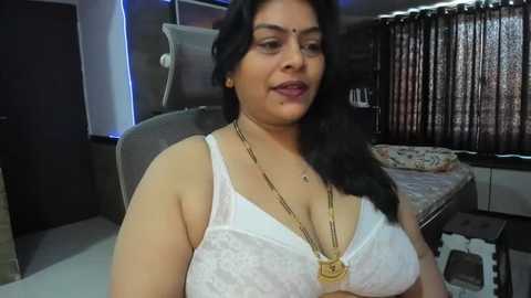 Media: Video of an Indian woman with medium skin tone, dark hair, wearing a white lace bra, gold necklace, sitting on a chair in a modern bedroom with patterned curtains and a bed.