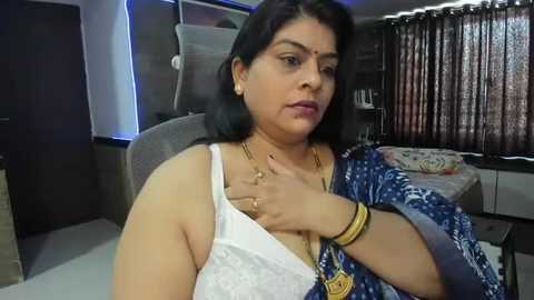 Media: Video of a South Asian woman with medium skin tone, medium build, wearing a blue saree, white blouse, and gold jewelry, sitting on a chair, looking serious.