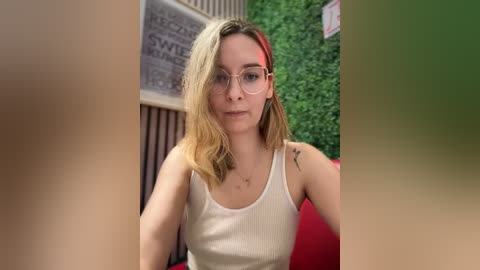 Media: Video of a young Caucasian woman with light skin, shoulder-length blonde hair, and round glasses, wearing a white tank top, seated in a modern caf\u00e9 with green foliage and red seating.