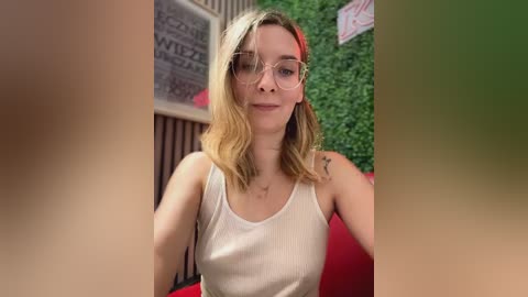 Media: Video of a young Caucasian woman with light skin and shoulder-length blonde hair, wearing wire-rimmed glasses and a white ribbed tank top, seated in a red chair with a green wall background.