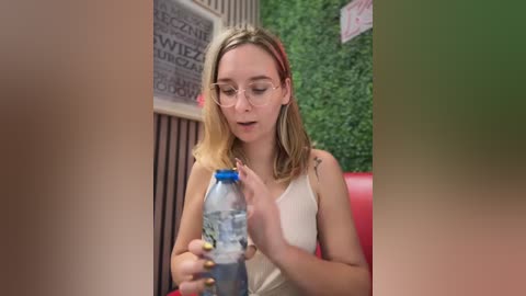 Media: A video of a fair-skinned woman with blonde hair, wearing glasses, drinking from a plastic bottle in a cafe with green walls and red chairs.