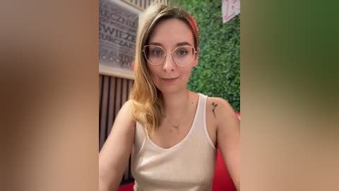 Media: A video of a young Caucasian woman with fair skin and blonde hair, wearing glasses, a white tank top, and sitting in a greenery-filled indoor setting.