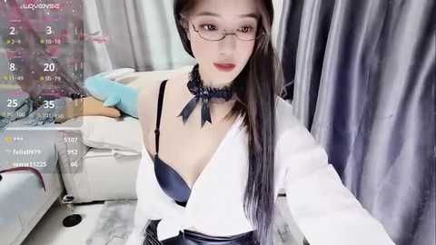 Media: Video of an Asian woman with long black hair, wearing glasses, a blue bow tie, and a revealing white blouse exposing her cleavage. She sits on a bed with pink and white sheets in a room with grey curtains.