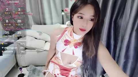 Media: A video of an Asian woman with long, straight black hair, wearing a revealing, floral-patterned dress, sitting on a white couch in a modern living room with gray curtains.