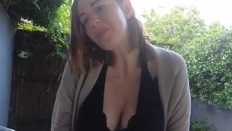 Media: Video of a fair-skinned woman with shoulder-length brown hair, wearing a black lace bra under a beige cardigan, smiling outdoors in a lush garden with green foliage and a wooden fence in the background.