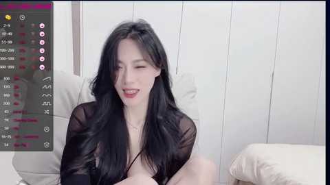 Media: Video of a smiling East Asian woman with long black hair, wearing a sheer black robe, sitting on a bed in a minimalist room.