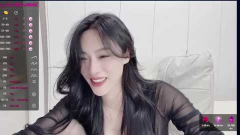 Media: A video of a young East Asian woman with long black hair, wearing a sheer black blouse, smiling in a modern, minimalist room with white walls and a white couch.